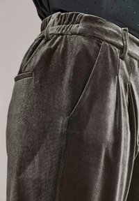 Close-up van donkergrijze fluwelen broek met zijzakken en een elastische tailleband, gedragen met een zwart blouse met stippen.