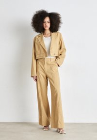 Blazer beige cropped avec deux boutons et pantalon beige à jambes larges. Le mannequin porte un top blanc et des talons de couleur claire. Texture de tissu lisse.