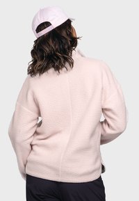 Heller rosa Fleece-Pullover mit strukturierter Oberfläche, hohem Kragen und überschnittenen Schultern, kombiniert mit einer weißen Mütze.