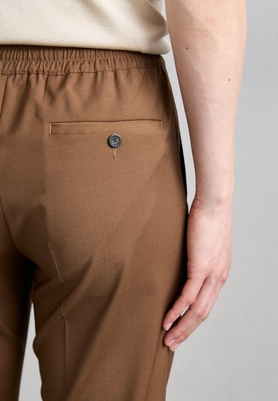 Brune bukser med elastisk talje, med en baglomme og en knaplukning. Stoffet har en glat tekstur med en subtil glans.