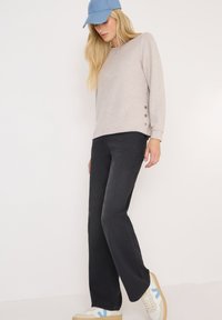 Sweatshirt beige clair avec des accents de boutons sur les côtés, pantalon noir évasé et baskets blanches avec des détails bleus, le tout surmonté d'une casquette bleue.