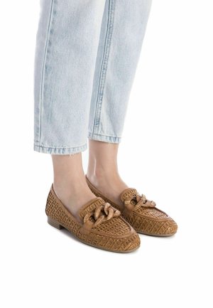 Mocasines tejidos en tono marrón claro con un acento anudado en el frente, que presentan un tacón bajo y plano y una superficie texturizada. Combinados con unos jeans cortos de color azul claro.