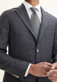 Giacca da abito grigia in lana con revers a punta, chiusura a un bottone e taschino sul petto. Abbinata a una cravatta grigia chiara e una camicia bianca.