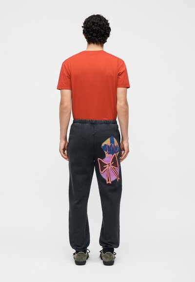 Άνδρας με κόκκινη μπλούζα και μαύρο παντελόνι φόρμας με πολύχρωμη φιογκάκι και γραφικά «1995» στέκεται γυρισμένος προς τα πίσω σε λευκό φόντο.