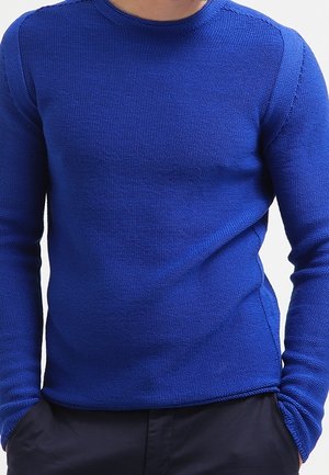 Mann trägt einen strukturierten, langärmligen, königsblauen Pullover mit den Händen teilweise in den Taschen einer dunklen Hose.