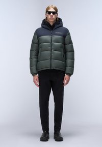 Green and navy puffer jacket with a zip front, features a detachable hood, elastic cuffs, and bold stitching. Paired with black trousers.