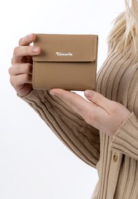 Hände halten eine kleine, hellbraune Lederbörse mit geprägtem Muster und dem "Tamaris"-Logo, getragen über einem beigen, gerippten Pullover mit Knopfdetail.