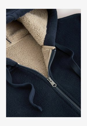Marineblaue Fleece-Zip-Hoodie mit beigem, weichem Futter und grauem Reißverschluss, ausgestattet mit einer Kapuze mit Kordelzug und strukturiertem Außenmaterial.