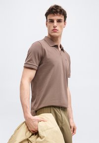 Άνδρας με καφέ πόλο μπλουζάκι και ελιά παντελόνι, κρατώντας μπεζ σακάκι στο αριστερό χέρι, στέκεται μπροστά σε απλό ανοιχτό φόντο.