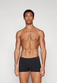 Muž s vypracovanou postavou stojaci čelom dopredu, oblečený v čiernych boxerkách s modrým logom "SLOGGI" na pásiku, na jednoliatom pozadí.