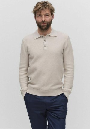 Beige knitted pullover with a collared neckline and three buttons, paired with dark navy trousers. Soft texture and snug fit.