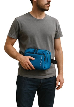 Riñonera azul de Nautica con dos compartimentos con cremallera, fabricada en un tejido suave y con una correa negra ajustable, sostenida por una persona que lleva una camiseta gris.