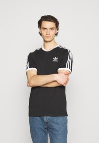 Črna majica s kratkimi rokavi z belim obodom in tremi belimi črtami na rokavih, z adidasovim logotipom na prsih. Ujemajo se z modrimi kavbojkami.