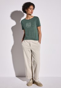 Grön kortärmad t-shirt med vit textdesign och krämfärgade videbyxor i kombination med beige sneakers.