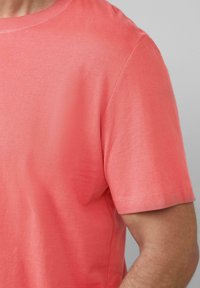 Kurzärmliges T-Shirt aus korallenfarbener Baumwolle mit klassischem Rundhalsausschnitt. Glatte Textur und schlichtes Design, ohne Muster oder zusätzliche Details.