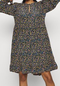 Femme portant une robe longue à manches longues, ample, avec un imprimé léopard multicolore et un petit détail en forme de trou de serrure au niveau du cou.