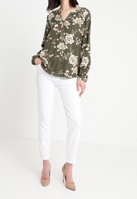 Kvinde iført en olivengrøn blomstret skjorte med lange ærmer, hvide slim-fit bukser med knapper ved anklerne og beige høje hæle, stående.