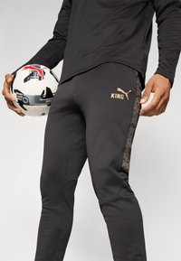 Melni sporta bikses ar zelta "KING" logo un rakstainu sānu svītru, kopā ar melnu un baltu futbola bumbu.