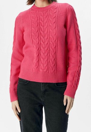 Frau trägt einen leuchtend pinkfarbenen Kabelstrickpullover mit strukturierten Mustern und schwarze Jeans, steht vor einem schlichten weißen Hintergrund.