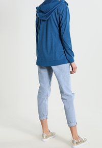 Blå huvtröja, mjukt tyg, avslappnad passform. Ljusblå jeans med upprullade ärmar, avslappnad stil. Guld sneakers med vita sulor.