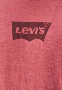 T-shirt de algodão vermelho com um logo da Levi's em castanho escuro, apresentando um design distinto em arco. Tecido suave com decote redondo e mangas curtas.
