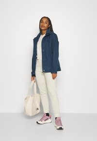 Marinfärgad vattentät jacka med dragkedjefickor, parat med ljusbeiga joggers, rosa sneakers och en krämfärgad tote-väska. Modellen står mot en enkel bakgrund.
