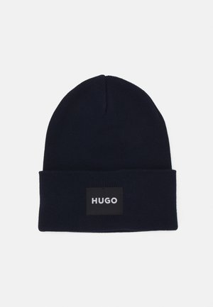 Marineblå hjemmestrikket hue med opsmøgget kant, der har en sort lap og "HUGO" i hvidt centreret på kanten.