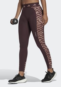 Leggings color bordeaux con un pannello a stampa zebra marrone, realizzati in tessuto elasticizzato, abbinati a scarpe da ginnastica nere e a un manubrio.