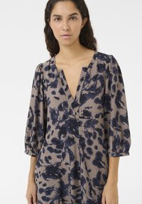 Robe marron avec des motifs abstraits bleu foncé, décolleté en V et manches bouffantes, fabriquée en tissu léger. Sontez un fit ample et des plis.