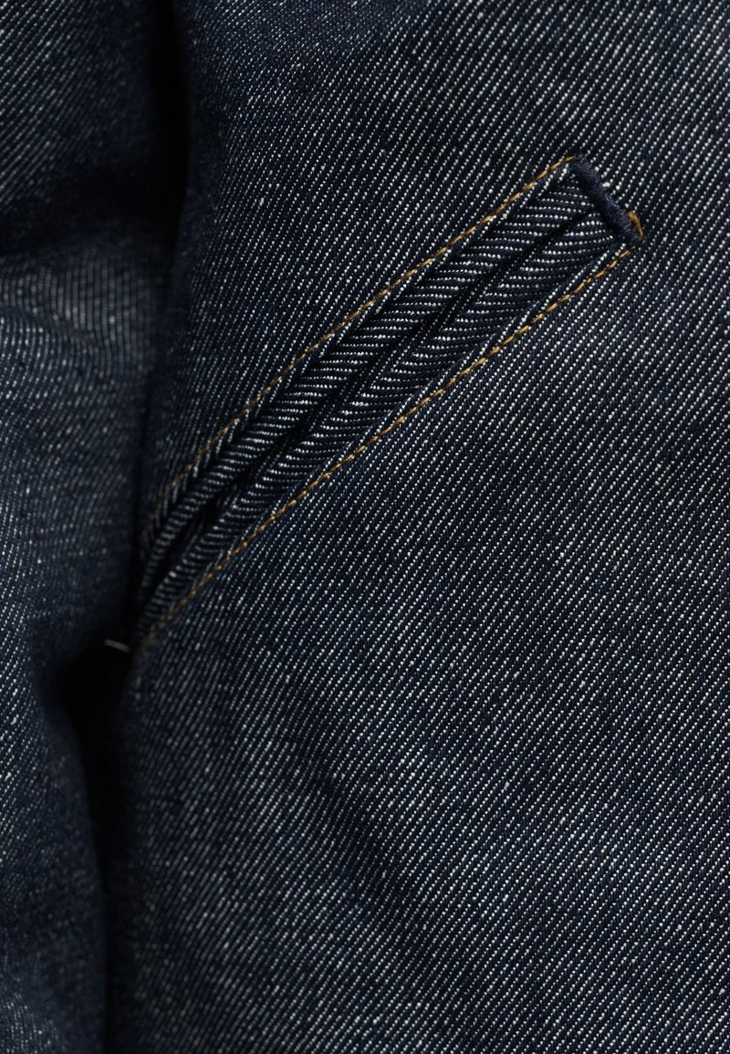 Tissu en denim foncé avec une texture subtile. Présente une vue rapprochée d'une ouverture de poche avec un design tressé le long de la couture de la fermeture éclair.