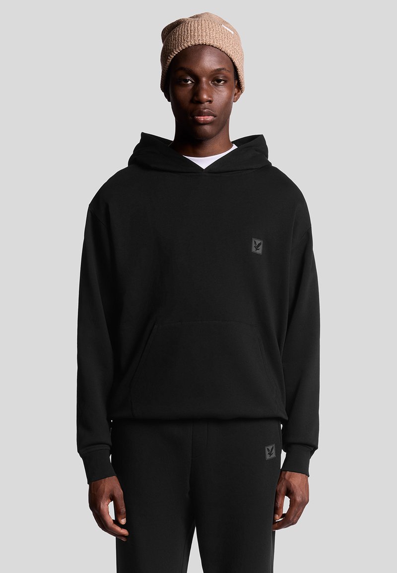 Un jeune homme portant un hoodie noir et un pantalon noir assorti, avec un bonnet en tricot beige, sur un fond gris uni.