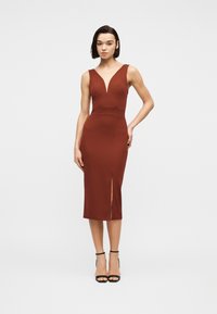 Γυναίκα που στέκεται φορώντας αμάνικο, μίντι φόρεμα σε σκουρόχρωμο πορτοκαλί απόχρωση με V-λαιμόκοψη και σκίσιμο μπροστά, συνδυασμένο με μαύρα πέδιλα με ψηλό τακούνι.