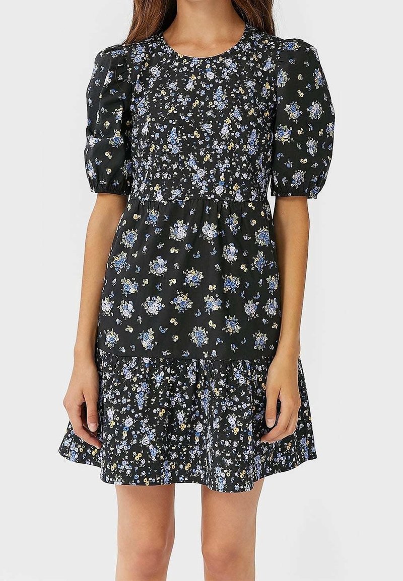 Femme portant une robe courte noire à fleurs avec des manches bouffantes et une jupe froncée avec des fleurs bleues et blanches.