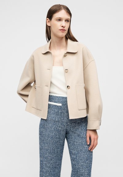 Γυναίκα που φοράει μπεζ σακάκι με κουμπιά και μεγάλες τσέπες, λευκό τοπ και μπλε υφασμένα παντελόνια με ψηλή μέση, στέκεται μπροστά σε ένα απλό φόντο.