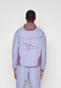 Άτομο με ανοιχτό μωβ και μπορντώ σακάκι και παντελόνι που ταιριάζει, με το κείμενο "ΜΗΝ ΤΡΕΧΕΙΣ ΑΠΛΑ" και ένα γραφικό παπουτσιού σε σχήμα αεροπλάνου στην πλάτη.