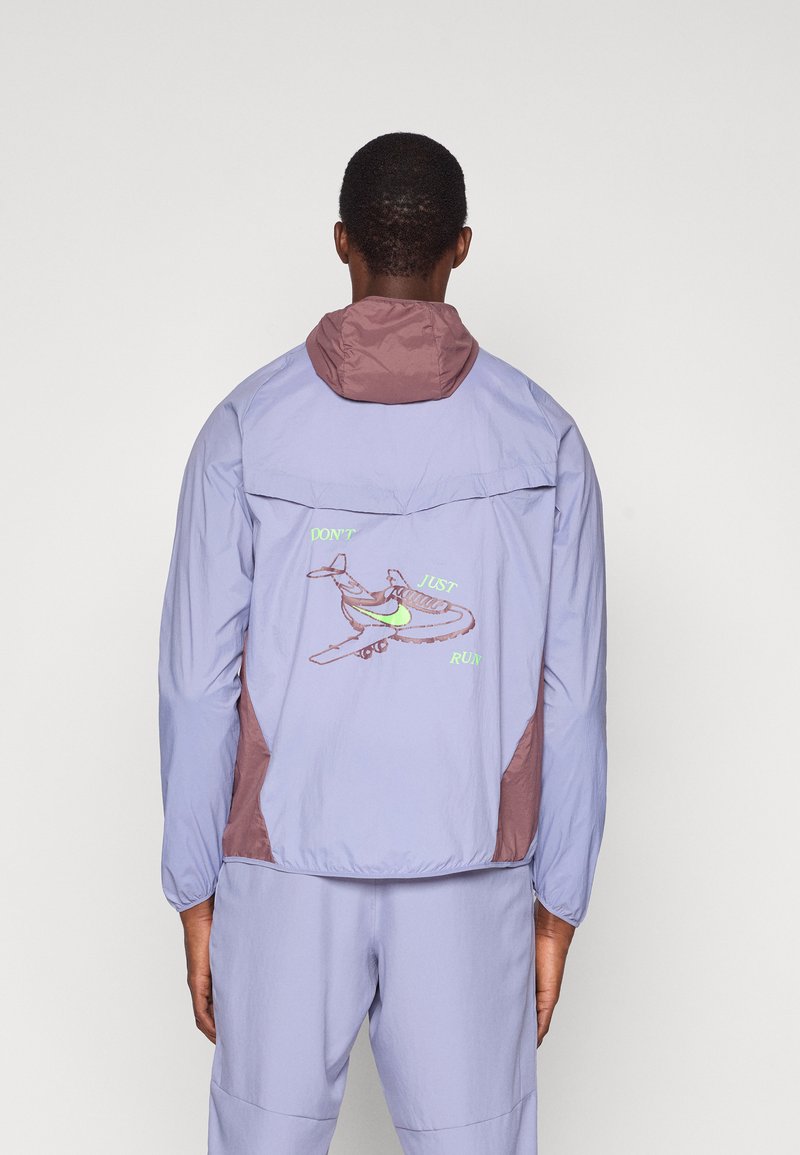 Άτομο με ανοιχτό μωβ και μπορντώ σακάκι και παντελόνι που ταιριάζει, με το κείμενο "ΜΗΝ ΤΡΕΧΕΙΣ ΑΠΛΑ" και ένα γραφικό παπουτσιού σε σχήμα αεροπλάνου στην πλάτη.