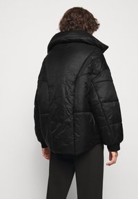 Osoba s kudrnatými vlasy, oblečená v černé pletené bundě a černých kalhotách, stojící zády k nám na jednobarevném světlém pozadí.