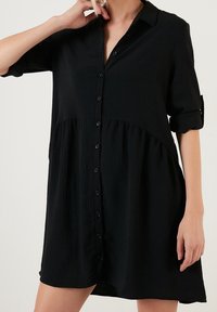 Abito nero a camicia con bottoni frontali, maniche arrotolabili, tessuto strutturato e parte inferiore svasata. Senza pattern né dettagli accentuati.