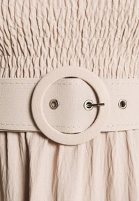 Světlé béžové plátno s texturovaným žabičkovaným designem. Široký pásek zdobí kruhová přezka a kovové průchodky, které zdůrazňují strukturované linie.