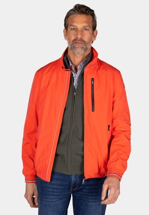 Rode lichtgewicht jas met een hoge kraag, ritszakken en gestreepte accenten. Draagt over een groene ritstrui. Blauwe jeans maken de look compleet.