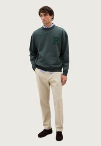 Felpa verde con un grande logo "P" verde sul petto, abbinata a pantaloni beige e scarpe marrone scuro. Camicia a righe blu visibile sotto.