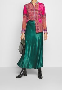 Bunte gemusterte Bluse mit Knöpfen, kombiniert mit einem grünen Satrock. Schwarze Knöchelstiefel und eine kleine strukturierte Handtasche runden den Look ab.
