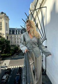 Šedý kardigan s výstřihem do V a velkými knoflíky, má volné rukávy. Nosený přes světlé, vzorované kalhoty. Pozadí zobrazuje městskou architekturu.