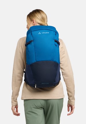 Zaino blu e blu scuro con più scomparti, spallacci regolabili e una texture liscia, etichettato "Skomer Zip 22" sul fondo.