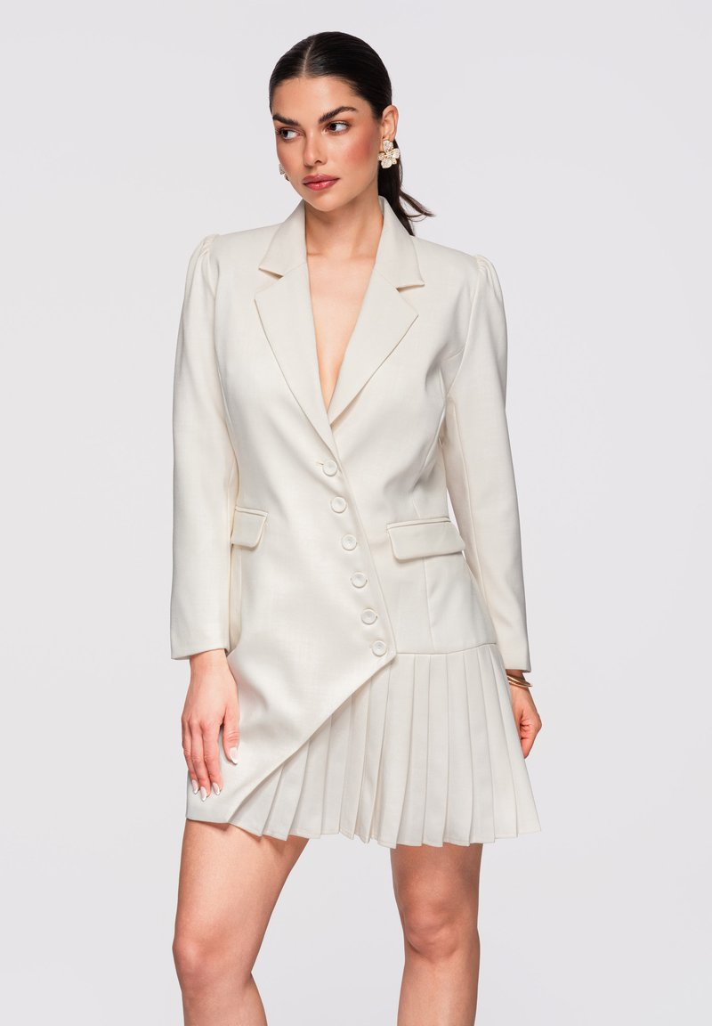 Femme aux cheveux foncés attachés en queue de cheval basse, portant une robe-blazer crème asymétrique avec une jupe plissée et des boucles d'oreilles fleuries, posant de profil.