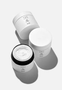 Trois pots cosmétiques. Deux blancs avec des surfaces lisses ; un noir avec une finition brillante. Un pot est ouvert, révélant un beurre corporel crémeux et blanc à l'intérieur.