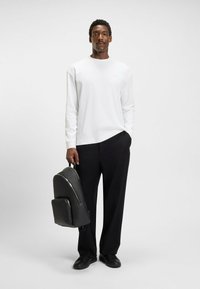 White long-sleeve shirt with a round neckline, paired with black trousers. The individual holds a textured black backpack with a front pocket.