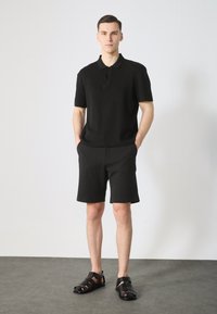 Polo de manga corta negro con tapeta de tres botones, combinado con pantalones cortos negros y sandalias negras con tiras. Textura suave, corte casual.
