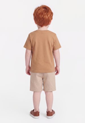 Jeune enfant aux cheveux roux portant un t-shirt marron, un short beige et des chaussures marron, debout de dos sur un fond blanc.