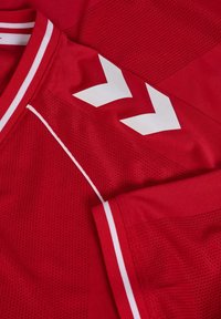 Maglia sportiva rossa con strisce a chevron bianche sulla spalla, sezioni in tessuto a rete e piping bianco lungo il colletto e i bordi delle maniche.